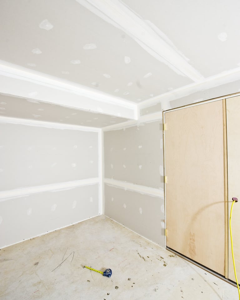 Room with concrete floor, drywall angles, bulkhead on ceiling, and studs marked on floor with tape measure