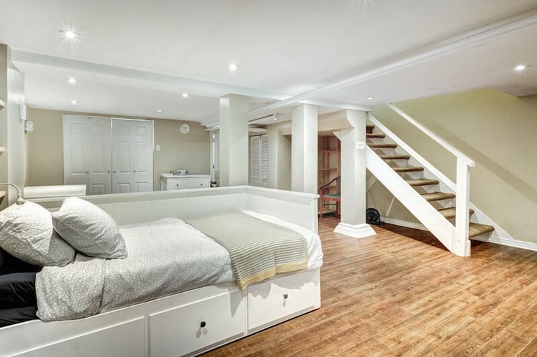 Modern basement bedroom with white platform bed, built-in storage, wooden flooring, and staircase leading upstairs