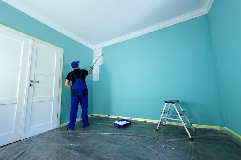 Painter in blue overalls applying turquoise paint to a room wall with a roller, ladder and paint tray visible on the floor
